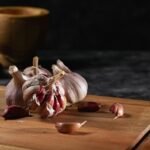 Close-up of garlic bulbs and cloves on a wooden cutting board with a rustic mortar in the background.