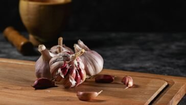 Close-up of garlic bulbs and cloves on a wooden cutting board with a rustic mortar in the background.