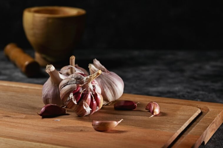 Close-up of garlic bulbs and cloves on a wooden cutting board with a rustic mortar in the background.