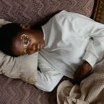 An adult woman relaxing indoors with artistic face paint, lying on a pillow and blanket in a cozy setting.