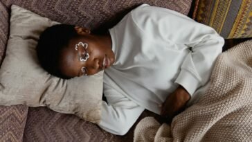 An adult woman relaxing indoors with artistic face paint, lying on a pillow and blanket in a cozy setting.