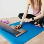 Woman in activewear using a laptop on a yoga mat with dumbbells, representing a healthy lifestyle.
