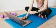 Woman in activewear using a laptop on a yoga mat with dumbbells, representing a healthy lifestyle.