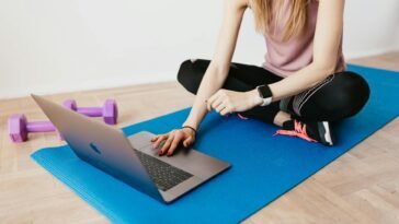 Woman in activewear using a laptop on a yoga mat with dumbbells, representing a healthy lifestyle.