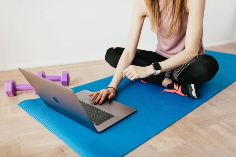 Woman in activewear using a laptop on a yoga mat with dumbbells, representing a healthy lifestyle.