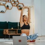 A woman in sportswear practicing yoga at home with a laptop nearby.