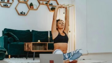 A woman in sportswear practicing yoga at home with a laptop nearby.