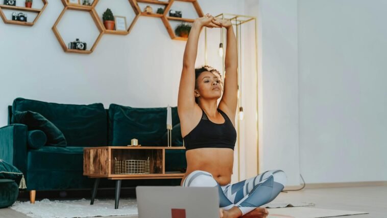 A woman in sportswear practicing yoga at home with a laptop nearby.