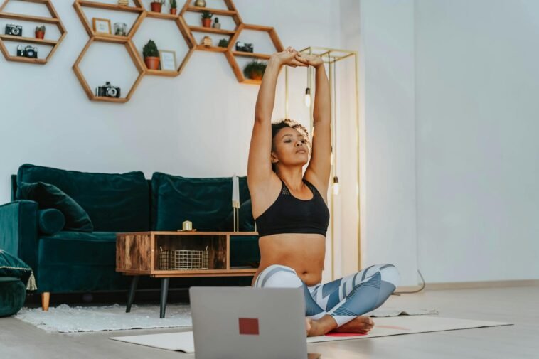 A woman in sportswear practicing yoga at home with a laptop nearby.