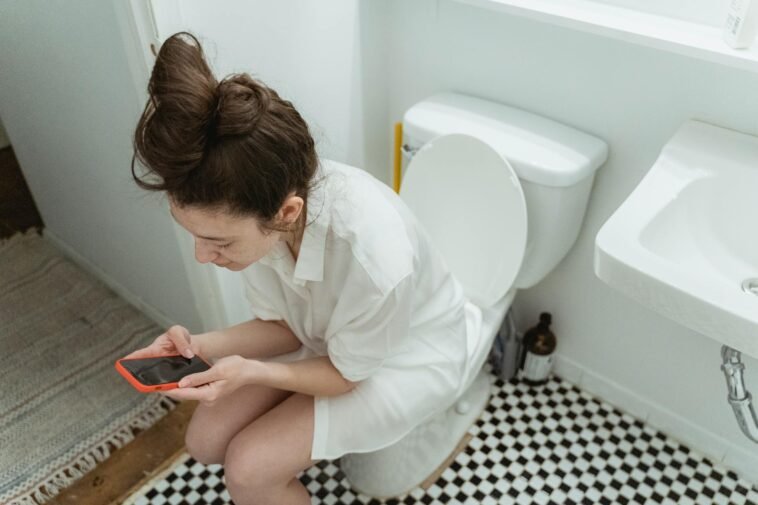 A woman sitting in a bathroom, focused on her smartphone, from a high angle view.