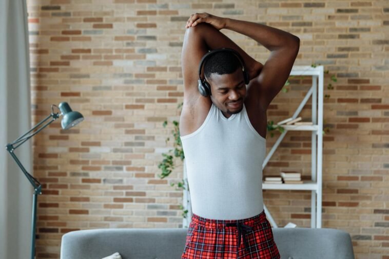 Man in white tank top stretching and listening to music indoors.