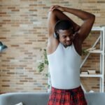 Man in white tank top stretching and listening to music indoors.