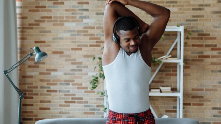 Man in white tank top stretching and listening to music indoors.