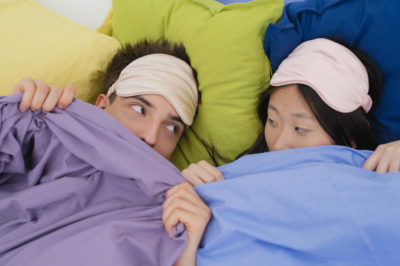 An interracial couple wearing sleep masks lying down on colorful pillows, portraying relaxation and rest.