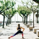 A woman in activewear performs a stretching routine on a sunny seaside walkway lined with trees.