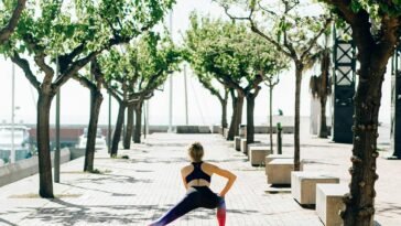 A woman in activewear performs a stretching routine on a sunny seaside walkway lined with trees.