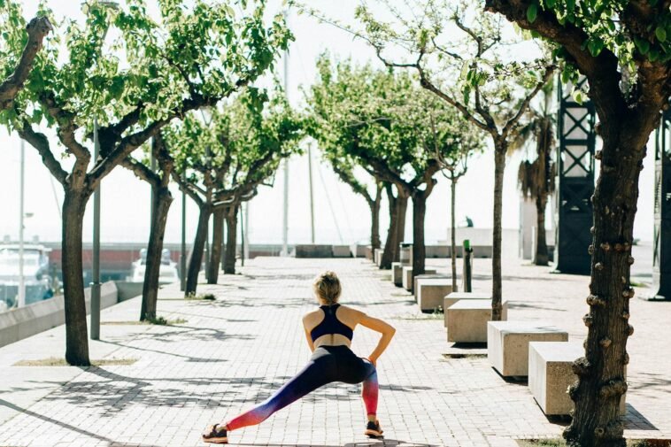 A woman in activewear performs a stretching routine on a sunny seaside walkway lined with trees.