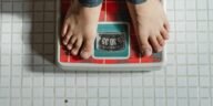 From above crop anonymous barefoot child in jeans standing on weigh scales on tiled floor of bathroom