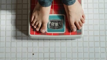 From above crop anonymous barefoot child in jeans standing on weigh scales on tiled floor of bathroom