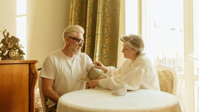 An elderly couple checks blood pressure using a digital monitor at their home table.